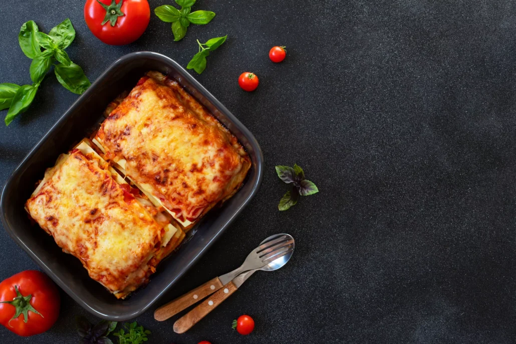 a freshly made lasagne in a black dish, on a black counter top with a fork, spoon, and scattered tomatoes