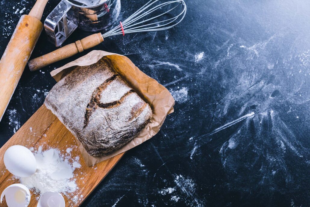 freshly baked loaf of bread wrapped in parchment paper on a black counter top with a whisk and a dusting of flour