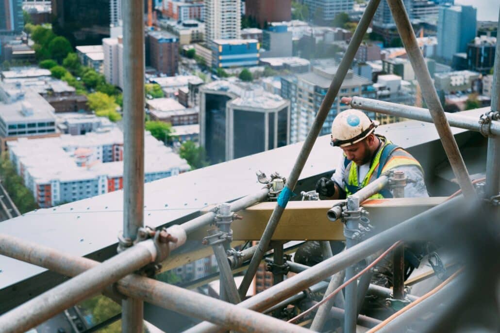 Construction worker wearing a safety harness and helmet working on scaffolding high above a city skyline with buildings in the background.